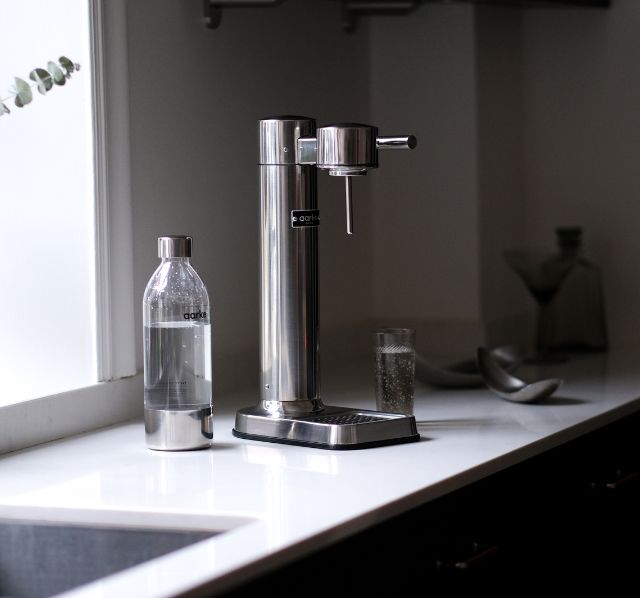 Ein Aarke Wassersprudler steht in einer minimalitischen Kuechenzeile, zusammen mit einer Aarke Wasserflasche und einem aufgefüllten Glas Wasser.