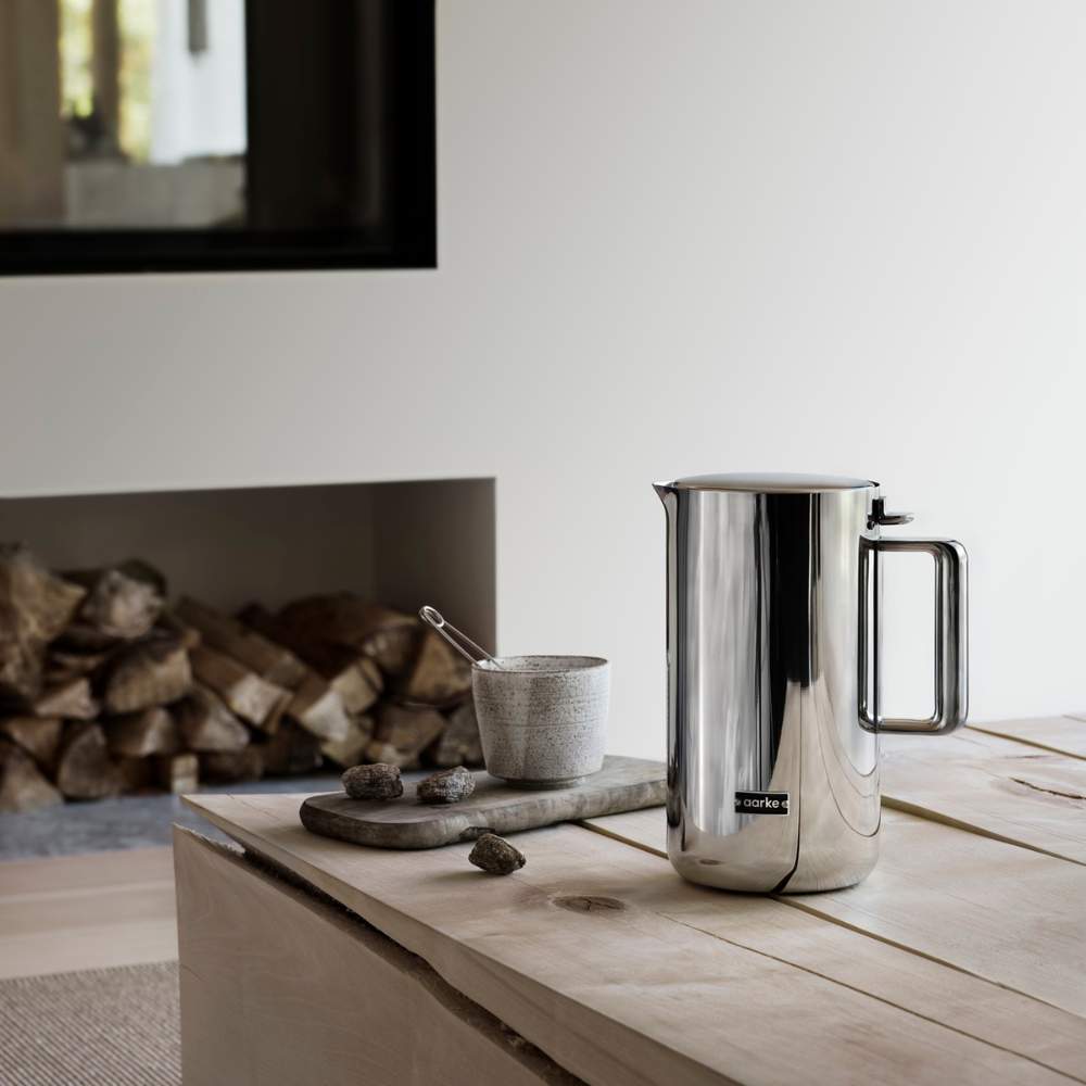 Ein Aarke Wasserkocher steht auf einem Holztisch vor einem offenen Kamin in einem modernen, lichtdurchfluteten Wohnzimmer.