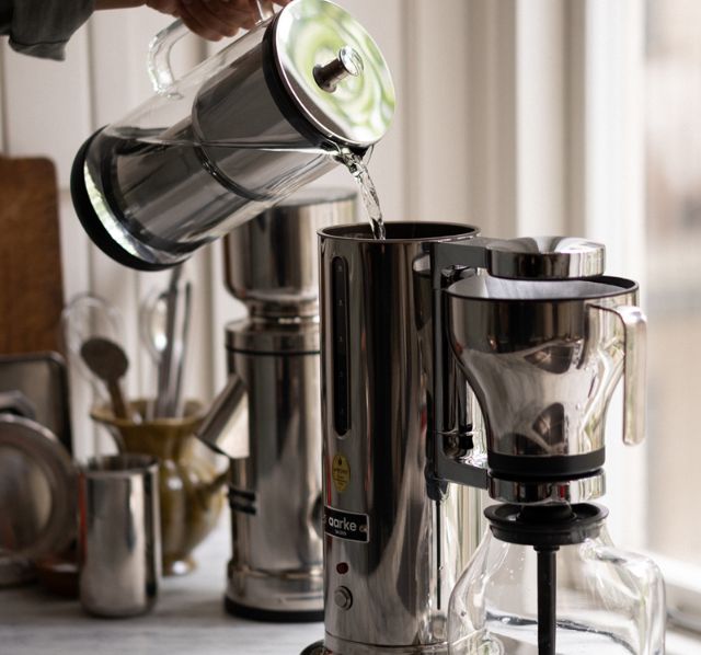 Eine Aarke Kaffeemaschine steht auf einer Marmor Kuechenplatte und wird mit frischem Wasser befüllt. Im Hintergrund sind verschiedene Kuechenutensilien zusehen.
