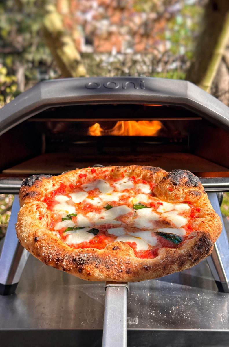 Eine neapolitanische Margherita Pizza wird angeschrägt auf einem Ooni Pizzaschieber präsentiert, während sie aus dem Ooni Koda Pizzaofen herausgenommen wird.