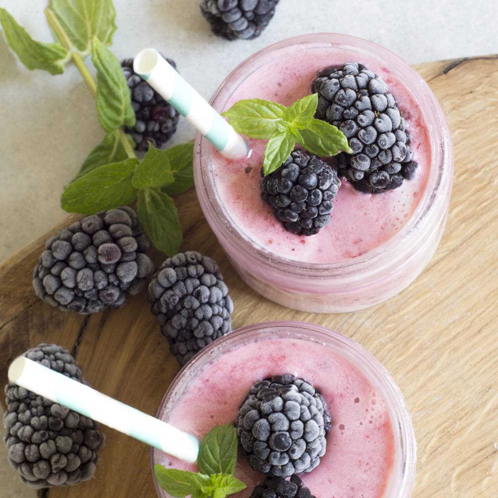 Zwei Smoothies aus Brombeeren mit Minzblättern dekoriert und jeweils zwei Strohhalmen stehen auf einem Holzbrett.