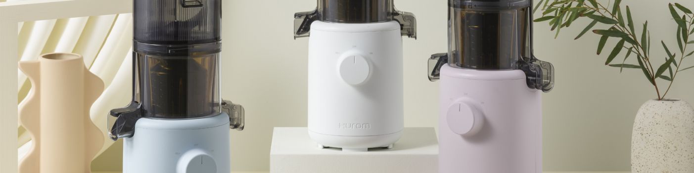 Drei Hurom Slow Juicer in Pastellfarben auf Podesten mit Pflanzen und Deko im Hintergrund.