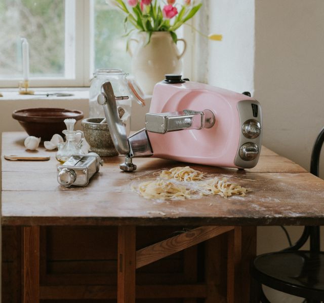 Rosa Ankarsrum Kuechengeraet mit Pastawalze steht auf einem rustikalen Holztisch. Neben dem Kuechengeraet liegen frisch gewalzte Nudeln.